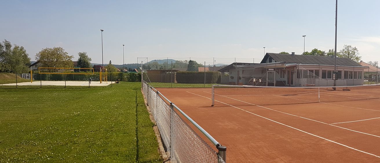 Ein leerer Tennisplatz mit braunem Boden und weißen Markierungen. Dahinter befindet sich ein Gebäude mit grauem Dach und Fenstern. Straßenlampen und Bäume im Hintergrund.