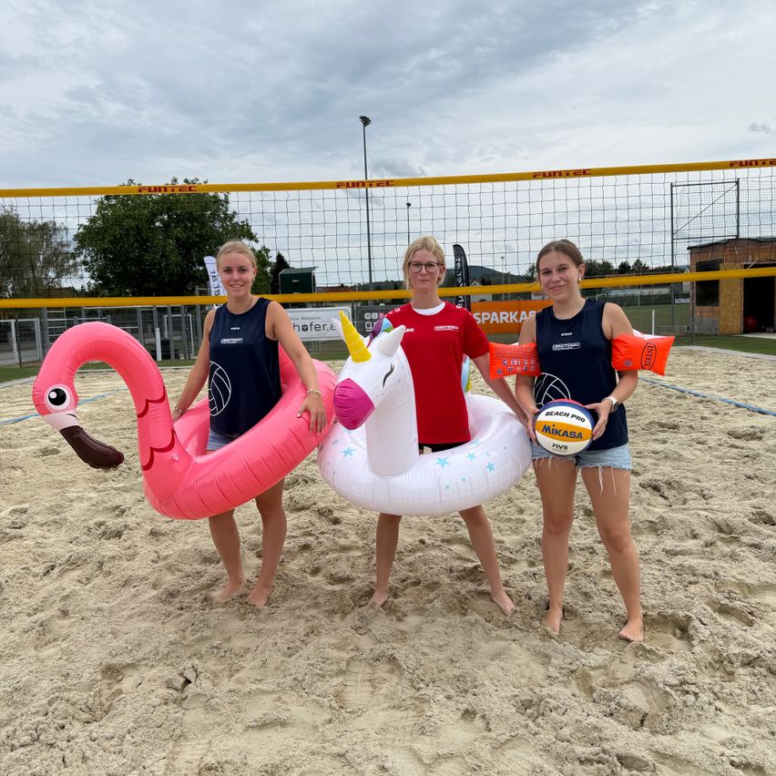 Drei Frauen stehen auf einem sandigen Strand und halten aufblasbare Schwimmtiere und einen Strandvolleyball. Sie lächeln und posieren für ein Foto. Hinter ihnen steht ein Volleyballnetz.