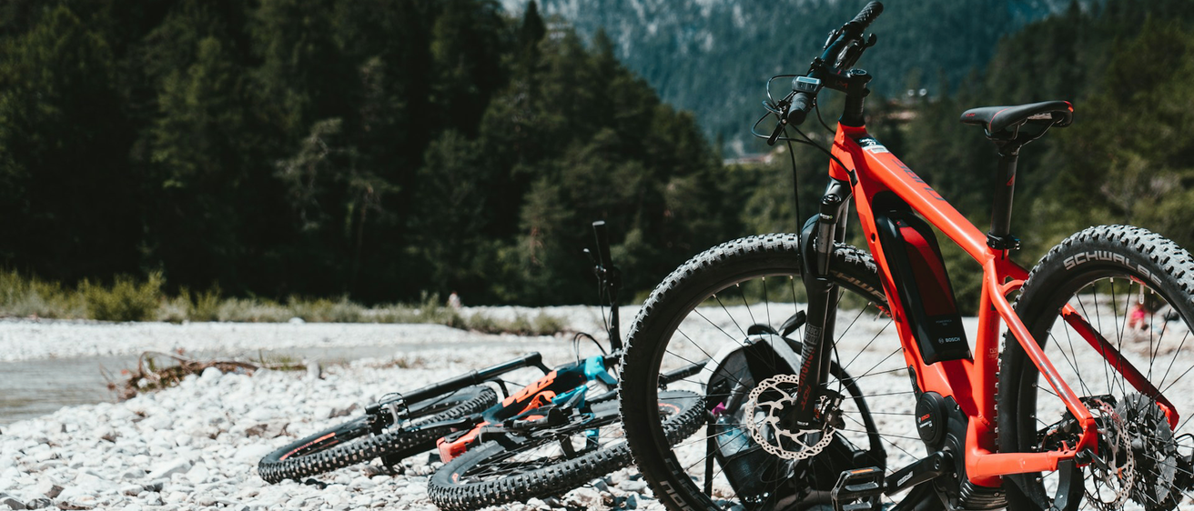 Zwei Mountainbikes sind auf einem Kiesweg mit bergiger Kulisse geparkt. Ein Fahrrad ist orange und das andere blau.