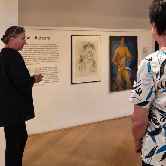 Eine Kunstgalerie mit einer Frau, die einer anderen Frau etwas erklärt, zwei Gemälde hängen an der Wand, eines ist ein Porträt und das andere eine nackte männliche Figur.
