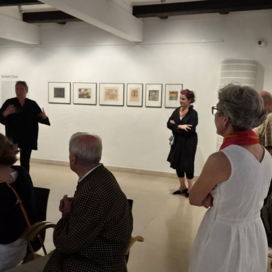 Eine Gruppe von Menschen ist in einem Raum, wahrscheinlich einer Kunstgalerie. Eine Frau steht und redet. Eine andere Frau steht mit verschränkten Armen. Der Raum hat eine weiße Wand mit Bilderrahmen.