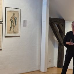 Eine Kunstgalerie mit Gemälden an der Wand. Eine Frau steht neben einem Holzbalken.