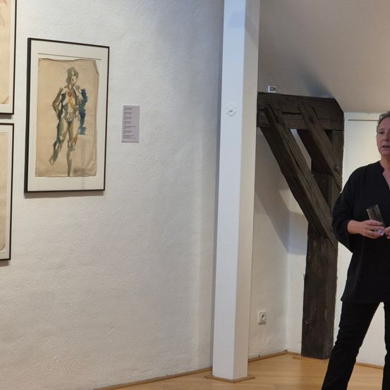Eine Kunstgalerie mit Gemälden an der Wand. Eine Frau steht neben einem Holzbalken.