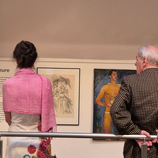 Eine Frau und ein älterer Mann betrachten Kunstwerke in einer Galerie. Die Frau trägt einen rosa Schal. Der Mann ist in einem karierten Anzug gekleidet.