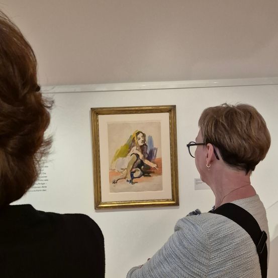 Zwei Frauen befinden sich in einer Kunstgalerie und betrachten ein Gemälde. Das Gemälde zeigt eine sitzende Frau mit einem gelben Schleier. Die Frau auf der rechten Seite trägt eine Brille und eine Halskette.