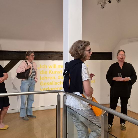 Eine Gruppe von Menschen steht in einem Innenraum, möglicherweise einer Kunstgalerie. Sie betrachten die Kunstwerke an der Wand. Die Frau vorne trägt eine Brille und eine Uhr. Die Frau rechts hält ein Glas. Die Frau links hält eine Handtasche. Der Boden ist aus Holz.