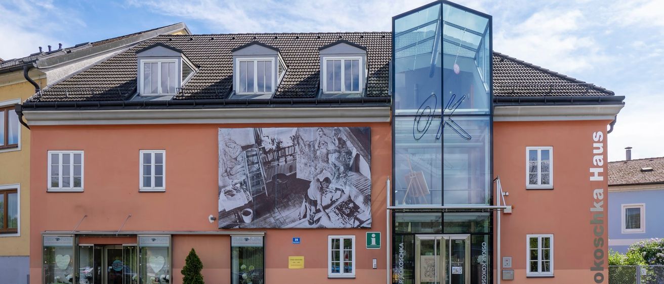 Die Vorderseite eines Gebäudes mit einem großen Wandbild, das Menschen bei der Arbeit darstellt. Es hat einen gläsernen Eingang mit den Buchstaben 'OK'.