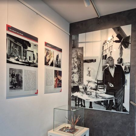 Ein Ausstellungsraum mit einem großen Schwarz-Weiß-Foto eines Mannes, der in einem Studio steht, verschiedenen kleineren Bildern an der Wand und einem Glaskasten mit Kunstzubehör.