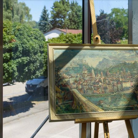Ein Kunstgemälde wird auf einem Staffelei vor einem Glasfenster ausgestellt, das eine Stadtlandschaft zeigt.