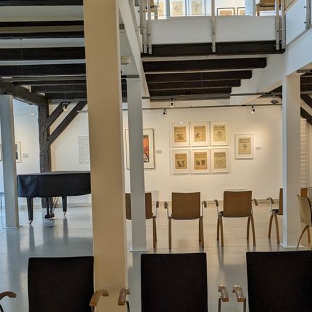 Ein Indoor-Kunstgalerie mit einem in schwarzen Stoff gehüllten Klavier. Kunstwerke sind an den weißen Wänden montiert, und mehrere Holzstühle sind angeordnet. Eine Treppe mit Handlauf ist rechts sichtbar.