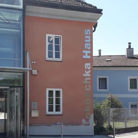Das Bild zeigt die Seite eines pfirsichfarbenen Gebäudes mit drei Fenstern und dem vertikal geschriebenen Text 'okoschcka Haus'. Rechts davon befindet sich ein blaues Gebäude mit zwei Fenstern, einem Schornstein und einem Zaun.