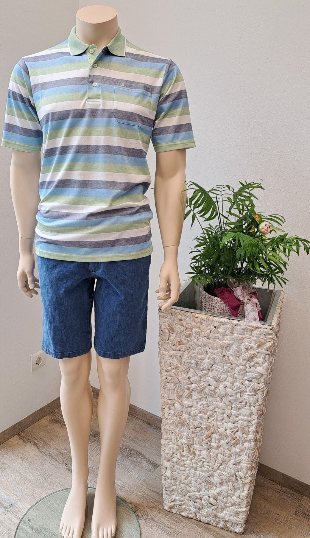 A mannequin displays a striped shirt and denim shorts. Next to it, there's a woven plant pot with a potted plant inside.