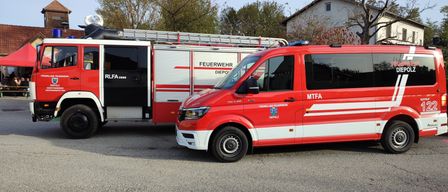 Ein roter Feuerwehrwagen und ein roter Van sind am Straßenrand geparkt. Der Feuerwehrwagen hat eine Leiter auf dem Dach und das Wort 'Feuerwehr' auf seiner Seite. Der Van hat das Wort 'MTFA' auf seiner Seite.