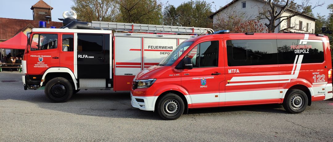 Ein roter Feuerwehrwagen und ein roter Van sind am Straßenrand geparkt. Der Feuerwehrwagen hat eine Leiter auf dem Dach und das Wort 'Feuerwehr' auf seiner Seite. Der Van hat das Wort 'MTFA' auf seiner Seite.