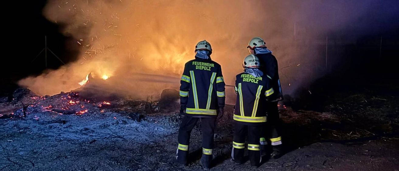 Drei Feuerwehrleute in Schutzkleidung stehen bei Nacht vor einem brennenden Feuer.