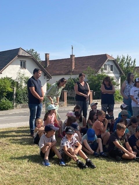 Eine Gruppe von Kindern und Erwachsenen sitzt auf dem Gras vor einer Reihe von Häusern. Erwachsene stehen hinter den Kindern.