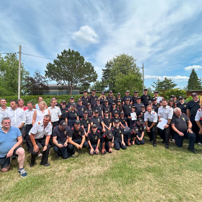 Eine Gruppe Feuerwehrleute, einige in Mützen und andere in Uniform, posieren für ein Foto auf einer Rasenfläche mit Bäumen und einem bewölkten Himmel im Hintergrund.