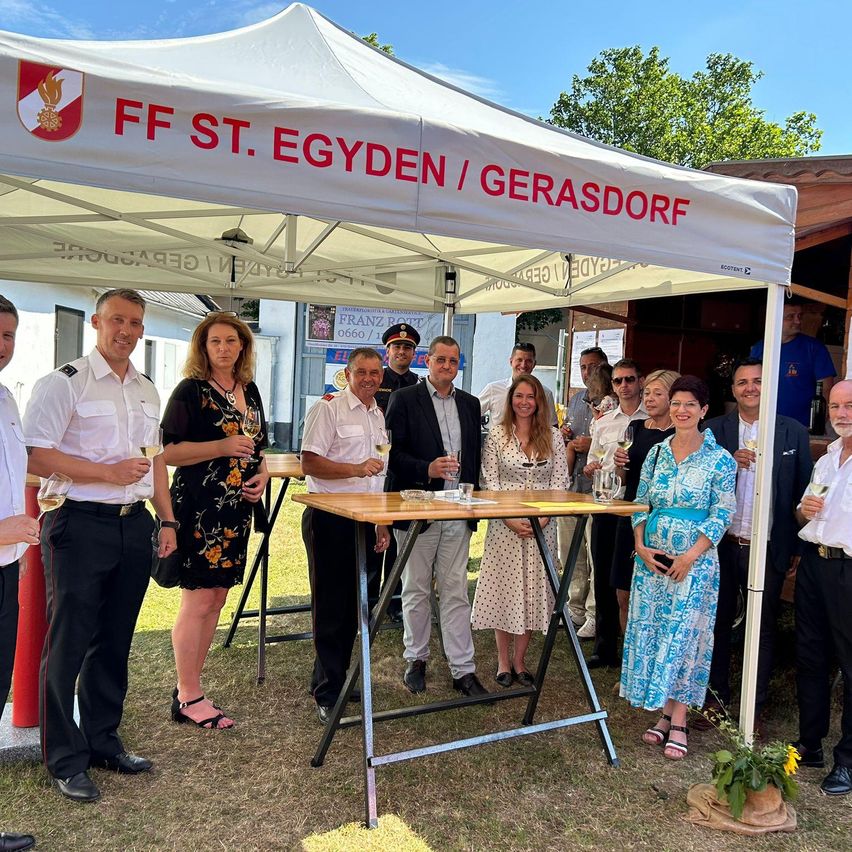 Eine Gruppe von Menschen steht unter einem Zelt mit der Aufschrift FF St. Egyden/Gerersdorf. Sie halten Weingläser in den Händen, einige in Uniformen, andere in Freizeitkleidung. Die Szene ist im Freien mit Tischen, einem Baum und einem Gebäude im Hintergrund.