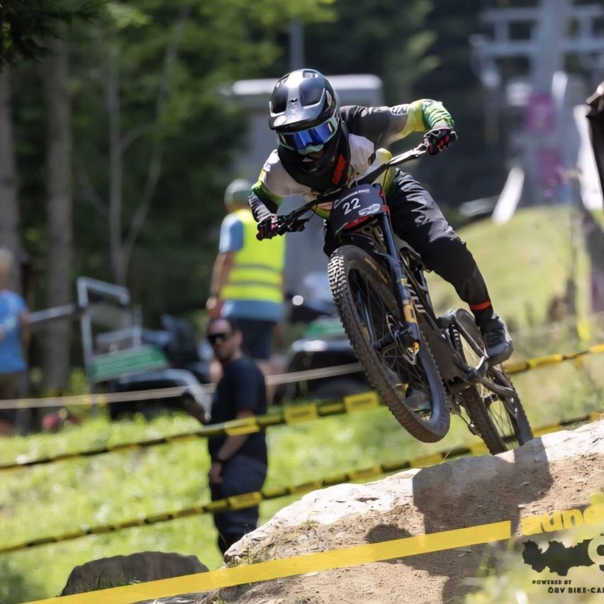 Ein Mountainbiker mit der Nummer 22 ist über einem Felshindernis in der Luft. Zuschauer und Mitarbeiter sind im Hintergrund.