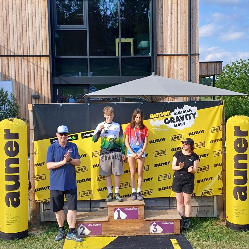 Drei Personen stehen auf einem Podium, eine auf dem ersten, eine auf dem zweiten und eine auf dem dritten Platz. Sie tragen Medaillen und stehen unter einem Banner mit 'Aunern Austrian Gravity Series'. Ein Mann klatscht hinter ihnen.