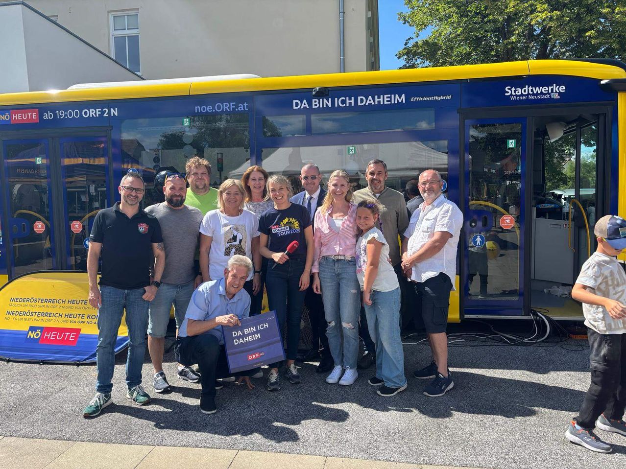 Eine Gruppe von Menschen steht vor einem blauen Bus mit dem Slogan DA BIN ICH DAHEIM. Der Bus ist auf der Straße geparkt.