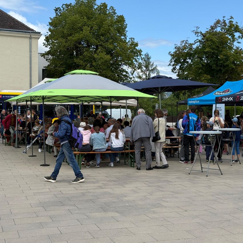Eine Gruppe von Menschen sitzt unter Schirmen bei einer Outdoor-Veranstaltung. Ein Mann mit Rucksack geht vorbei und entfernt sich von der Menge.