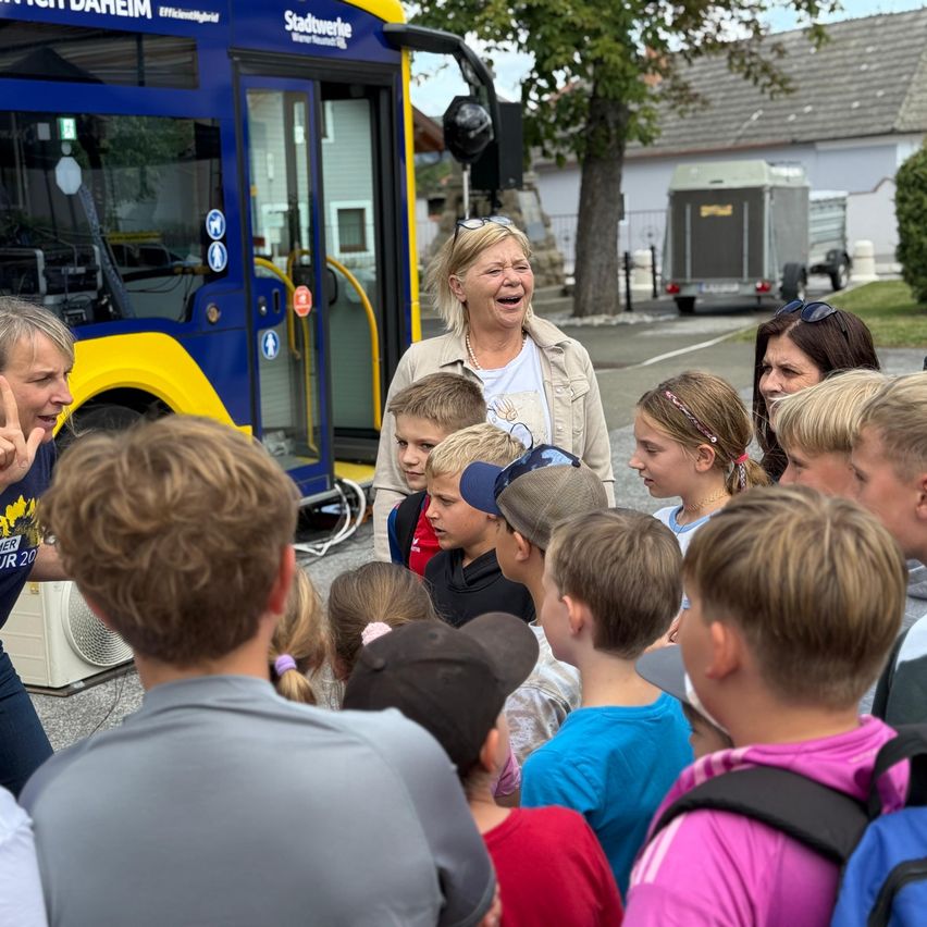 Eine Frau steht vor einem blauen Bus, lächelt und mehrere Kinder haben sich um sie versammelt. Ein Baum und ein Haus sind im Hintergrund zu sehen.