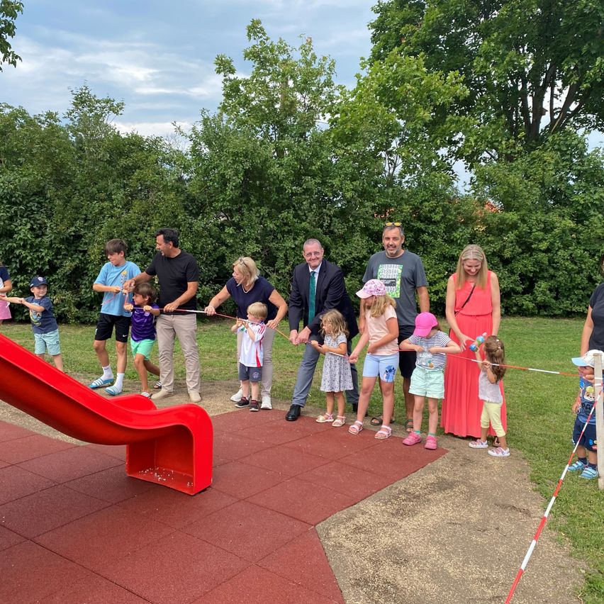 Eine Gruppe von Erwachsenen und Kindern steht in einem Park und hält ein Band, sie scheinen es zu schneiden. Es gibt einen roten Schlitten, Bäume und einen Rasenbereich.