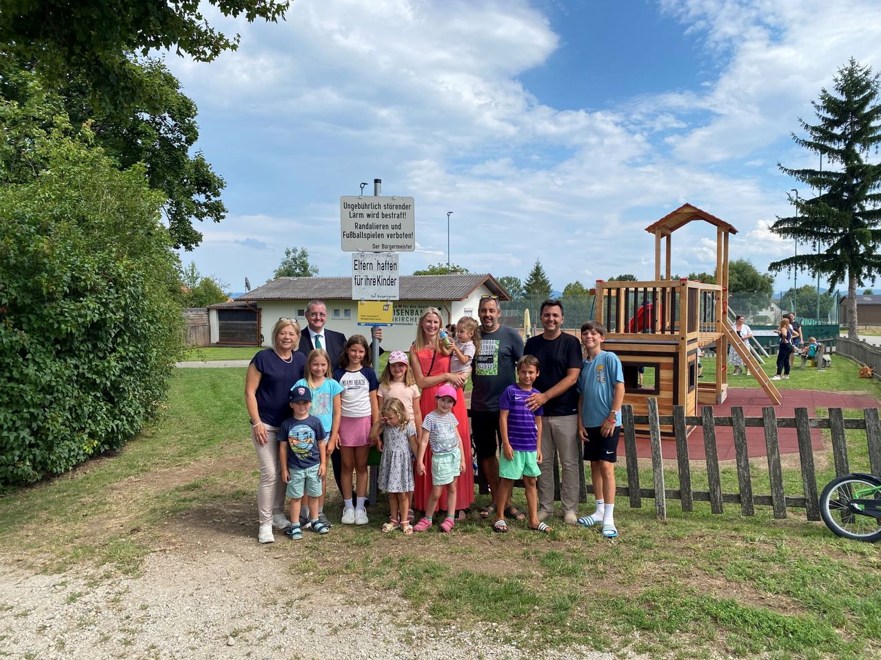 Eine Familie und mehrere Kinder posieren vor einem Spielplatz. Sie sind lässig gekleidet, und die Erwachsenen lächeln. Ein Schild hinter ihnen lautet 'Ungehublich sterben'.