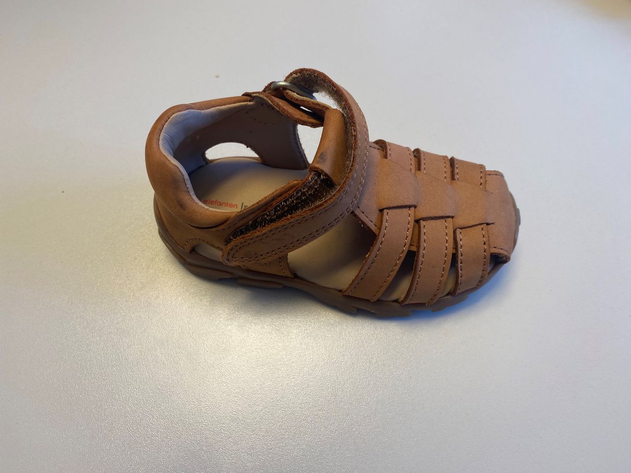 Ein brauner Ledersandal mit mehreren Riemen und Schnallen liegt auf einer weißen Oberfläche. Der Riemen am Zeh ist braun und die anderen Riemen sind beige.