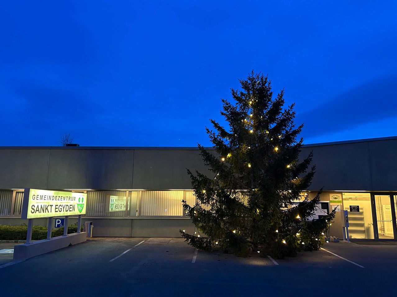 Ein geschmückter Weihnachtsbaum steht auf dem Parkplatz eines Gebäudes. Der Baum ist mit bunten Lichtern beleuchtet. Ein Schild am Gebäude lautet "Zentrum".