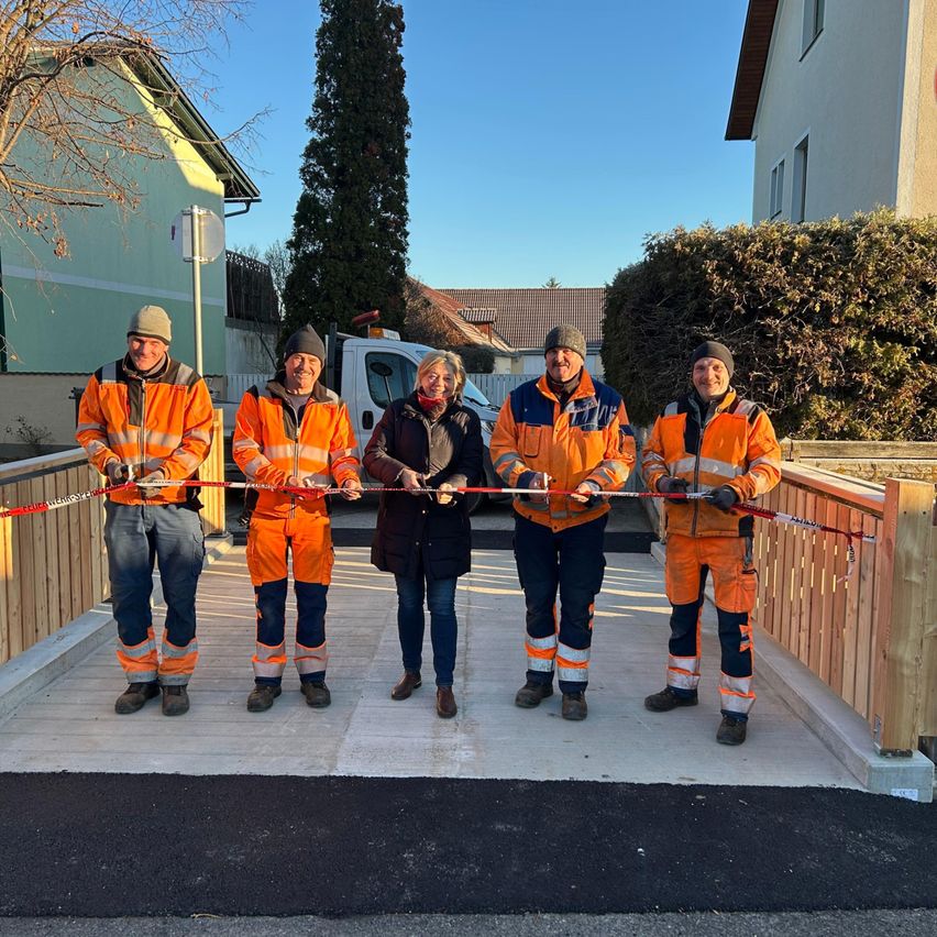 Fünf Personen in Sicherheitsausrüstung schneiden ein Band auf einer neu errichteten Brücke. Sie stehen vor Häusern und einem weißen LKW.