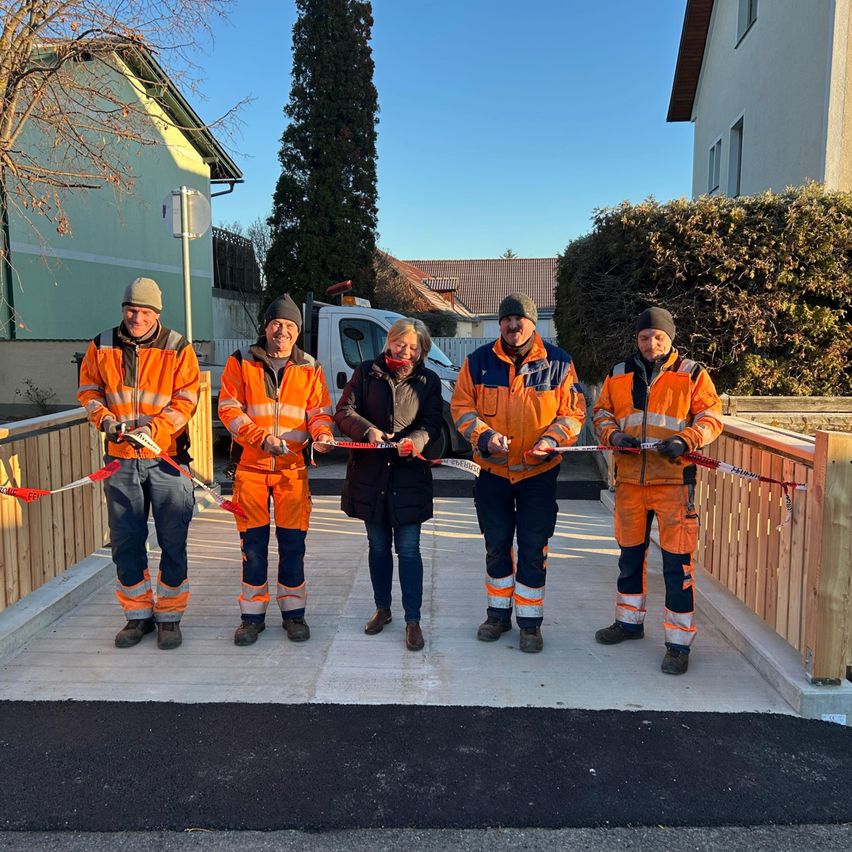 Fünf Personen in Warnwesten stehen auf einem neu gebauten Holzweg und halten ein rotes Band, wahrscheinlich bei einer Eröffnungsfeier. Dahinter stehen zwei Häuser, eines mit einem Baum, und ein weißer LKW.