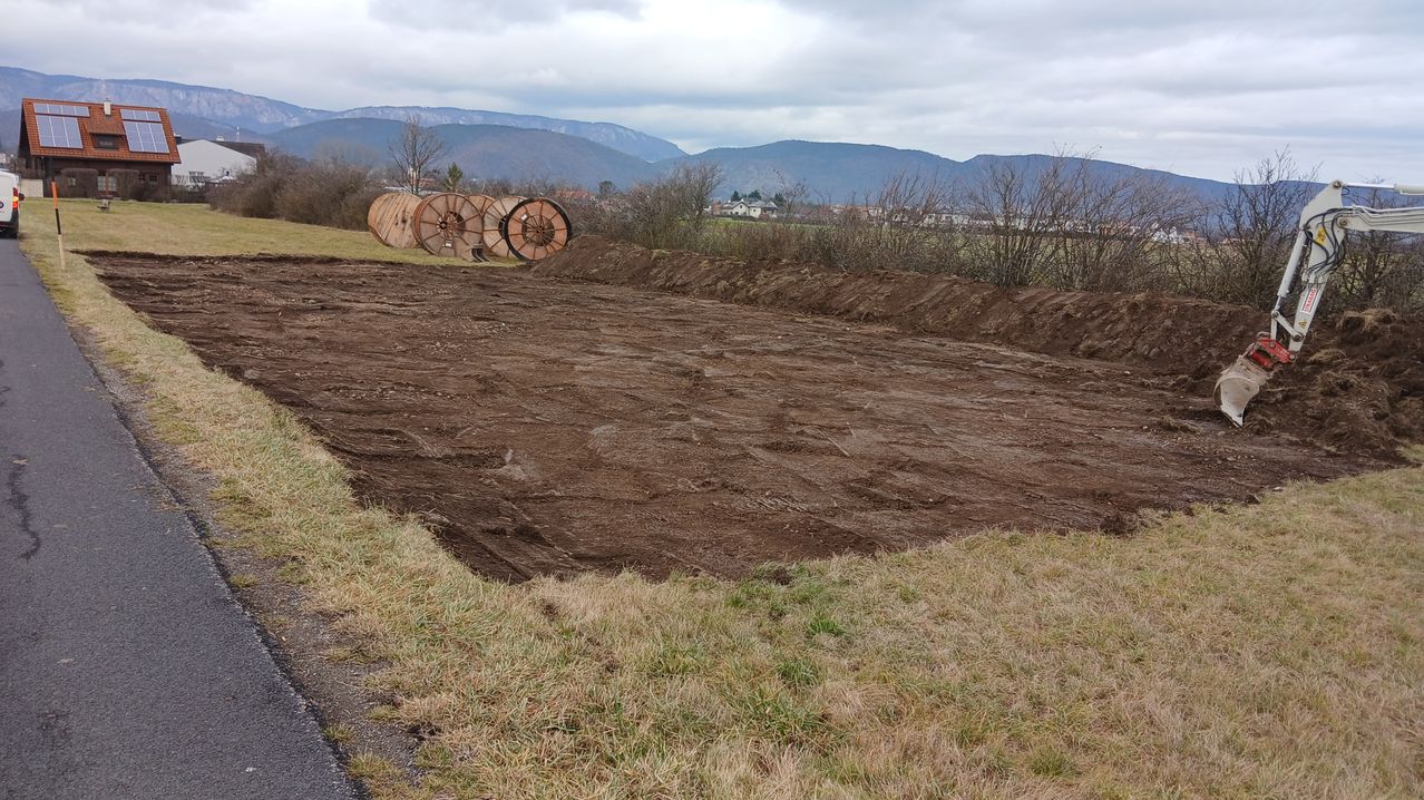 Ein Außenbereich mit großen, braunen Holzrädern an der Seite und einem großen, offenen, braunen Erdfeld. Im Hintergrund befinden sich Berge.