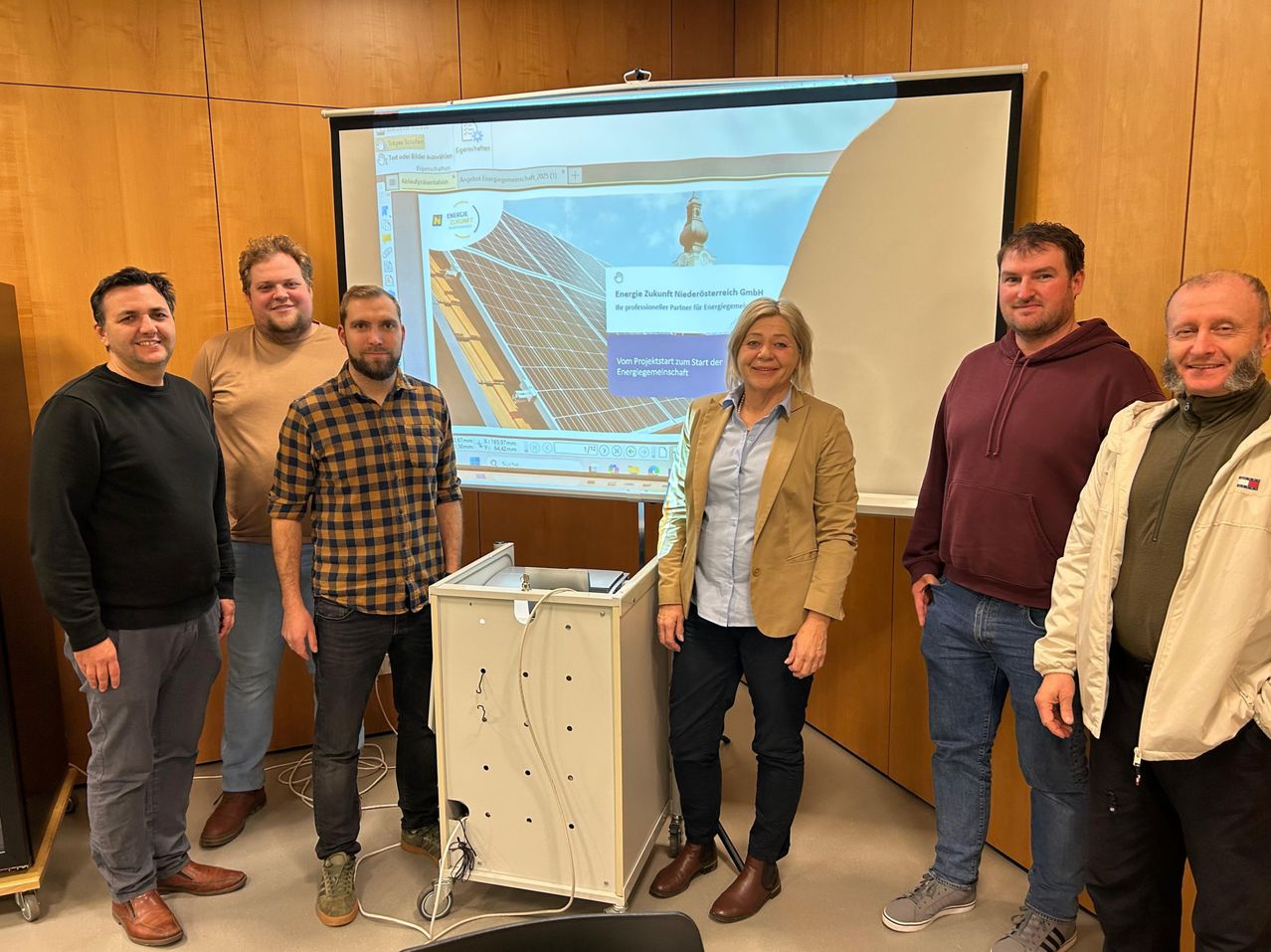 Eine Gruppe von Menschen steht neben einem Monitor in einem Raum, möglicherweise für eine Präsentation. Eine Frau in einer beigen Jacke steht vor einem Computer.