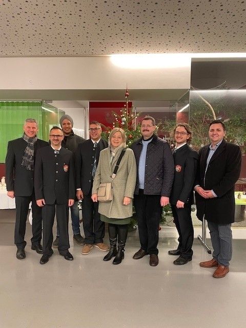 Eine Gruppe von Menschen steht vor einem Weihnachtsbaum, gekleidet in formelle Kleidung. Die Frau in der Mitte trägt eine Handtasche. Hinter ihnen ist ein Tisch aufgebaut mit einem Weihnachtsbaum und einem grünen Vorhang.