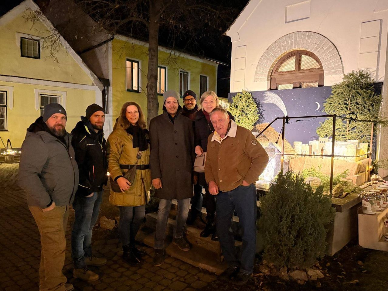 Eine Gruppe von Menschen steht nachts vor einem Haus. Sie lächeln und scheinen für ein Foto zu posieren. Vor ihnen stehen ein Baum und eine Pflanze.