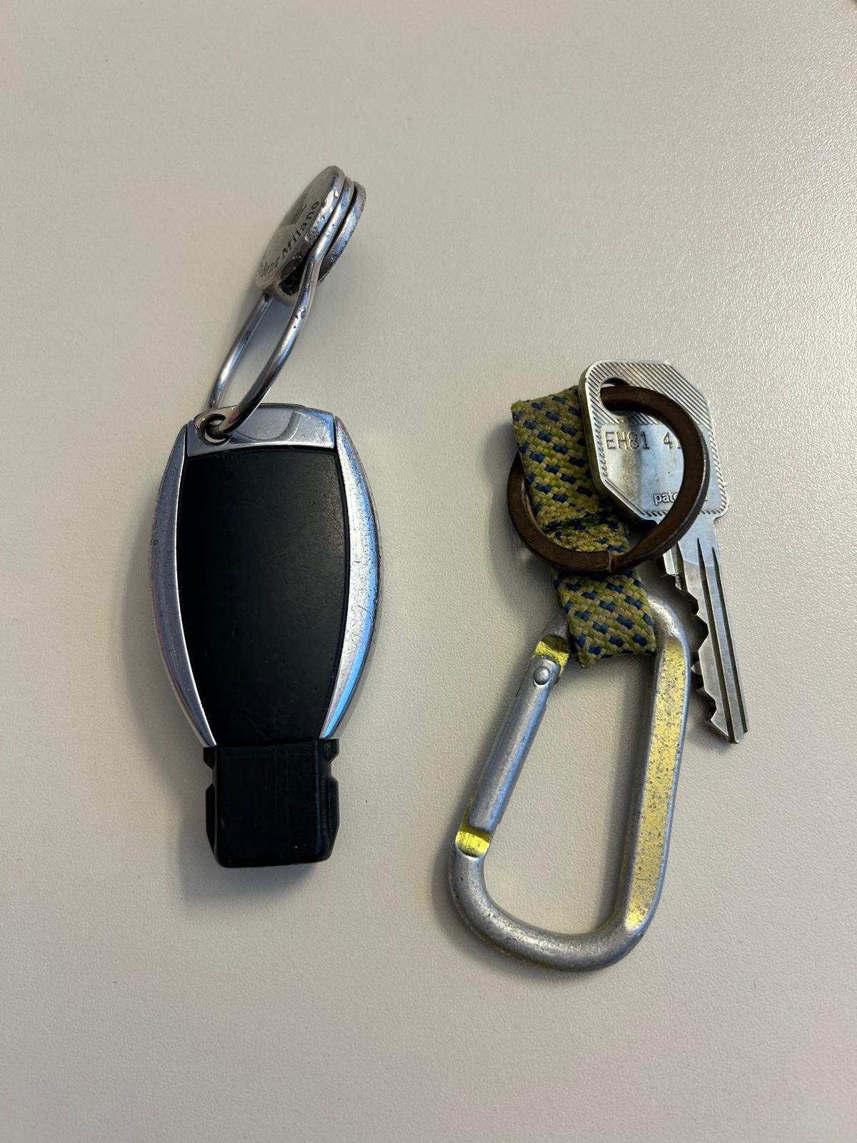 Zwei Schlüssel sind auf einer weißen Oberfläche zu sehen. Ein Schlüssel hat einen silbernen Karabiner mit einem gelben und grünen Gurt, während der andere Schlüssel einen schwarzen und silbernen Schlüsselbund mit den Buchstaben "OHM" darauf hat.