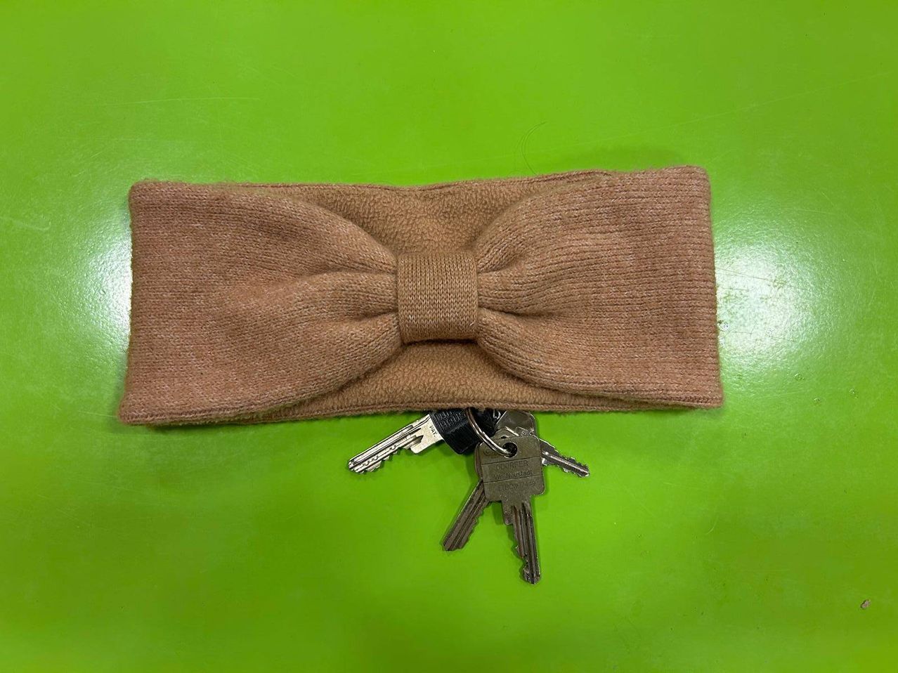 Ein braune gestrickte Stirnband mit einer Schleife liegt auf einer grünen Oberfläche und wird von einem Schlüsselbund begleitet.
