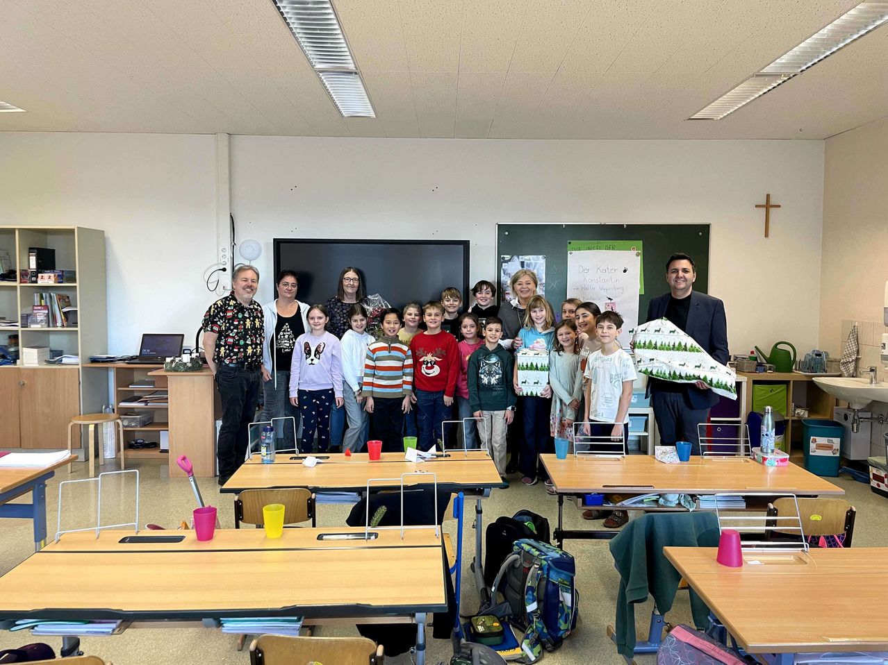 Eine Gruppe von Kindern und Erwachsenen steht in einem Klassenzimmer. Zwei Erwachsene halten ein großes Schild. Das Klassenzimmer hat Tische, eine Tafel und ein Kreuz an der Wand.