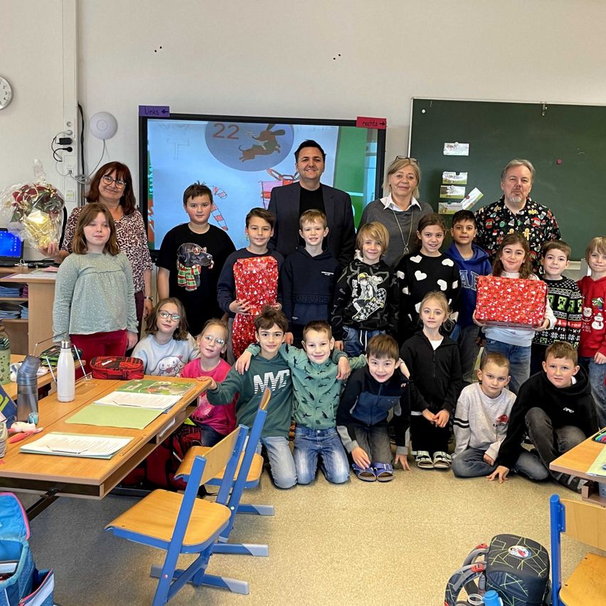 Eine Gruppe von Kindern und Erwachsenen posiert für ein Foto in einem Klassenzimmer mit einer großen Leinwand, Lehrern und verschiedenen Klassenzimmergegenständen.