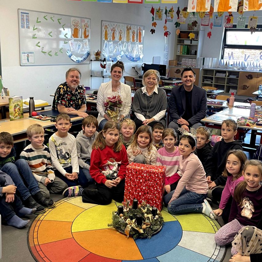 Gruppenfoto von Erwachsenen und Kindern in einem Klassenzimmer. Ein bunter Kreis auf dem Boden hat eine Girlande und Kerzen. Eine große Geschenkbox steht in der Mitte.