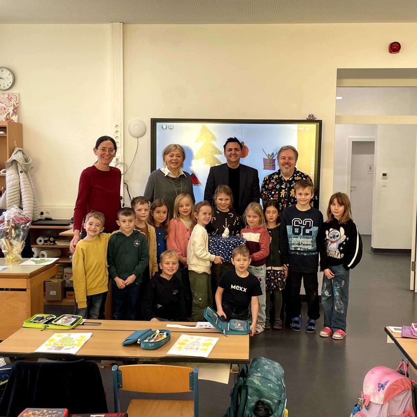 Eine Gruppe von Kindern und Erwachsenen posiert für ein Foto in einem Klassenzimmer mit einem großen Bildschirm im Hintergrund, der einen Weihnachtsbaum zeigt.