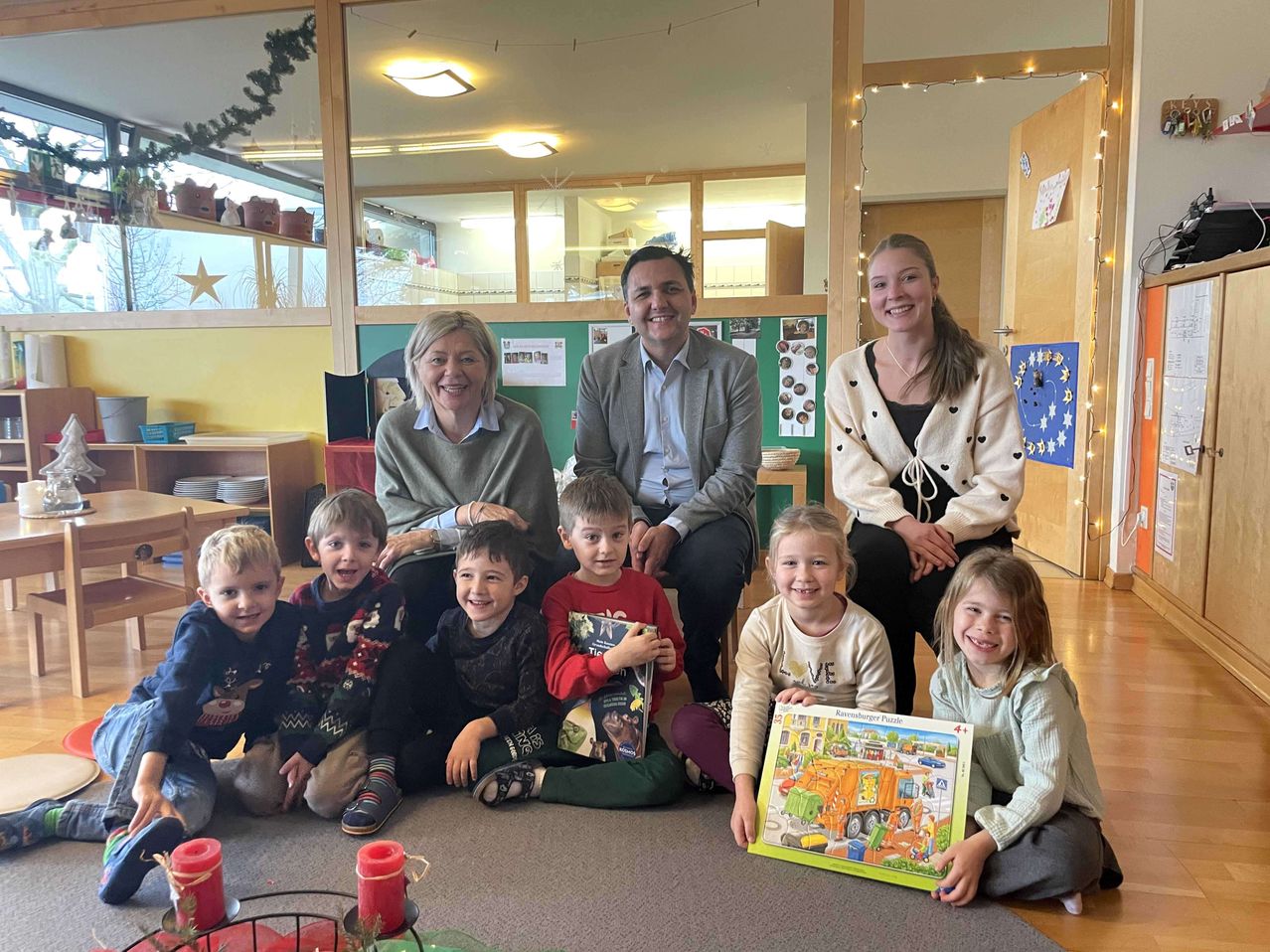 Mehrere Erwachsene und Kinder posieren in einem Klassenzimmer mit Teppich, Kerzen und Dekorationen. Einige Kinder halten Bücher und Puzzles.