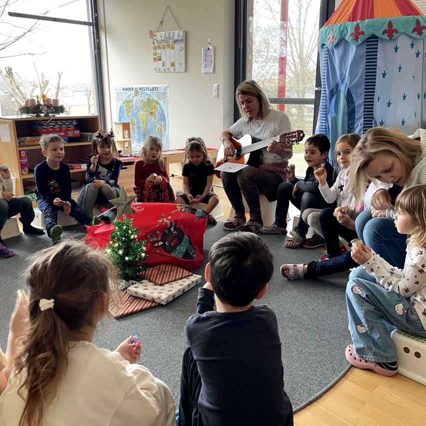 Eine Frau spielt Gitarre für eine Gruppe von Kindern, die im Kreis sitzen. Einige Kinder tragen Weihnachtsdekorationen und Geschenke liegen auf dem Boden. An der Wand befinden sich Bücher und eine Karte.