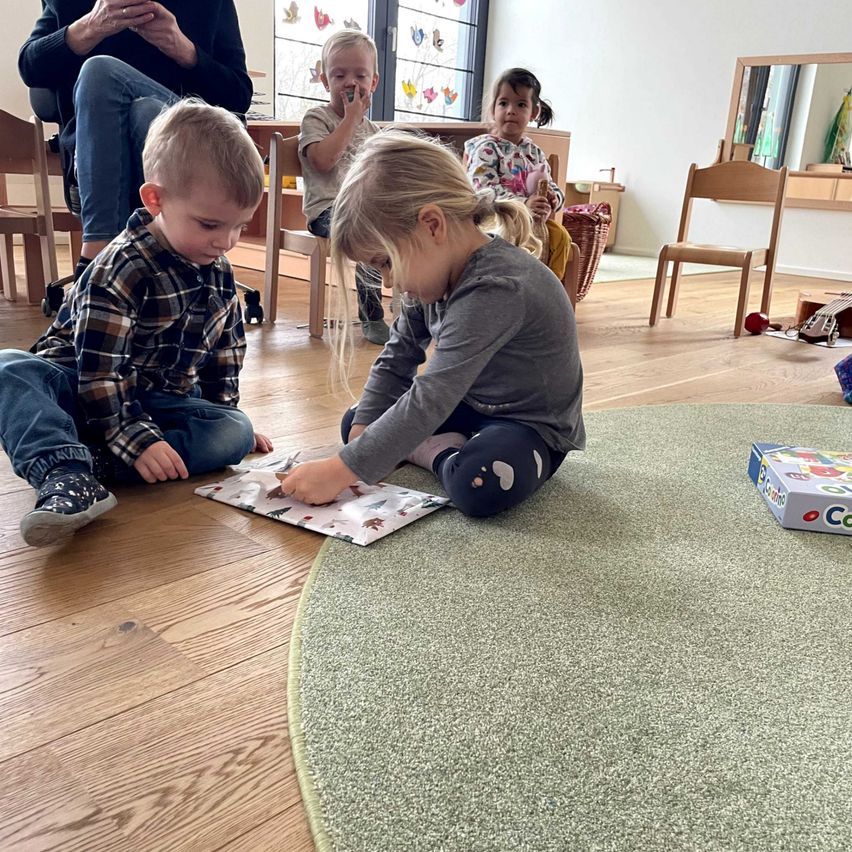 Eine Gruppe junger Kinder spielt auf dem Boden in einem Raum. Ein Mädchen öffnet ein Geschenk, während ein Junge zuschaut. Hinter ihnen sitzt eine Frau auf einem Stuhl. Es gibt einen Korb und einen Stuhl im Raum.