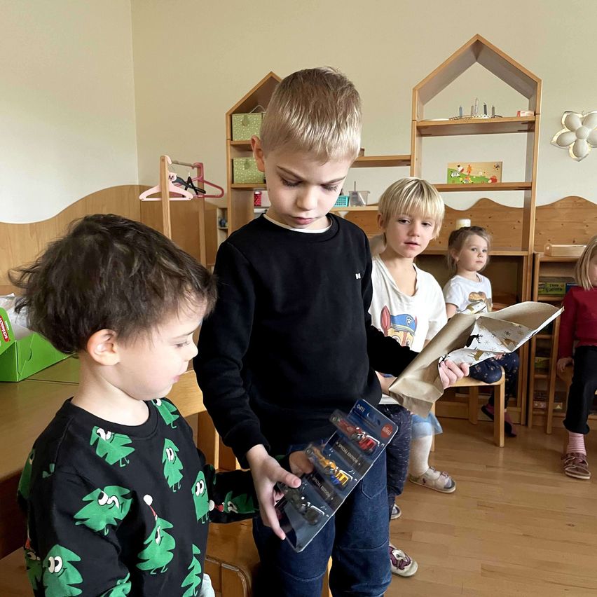 Zwei junge Jungen, einer mit einem Paket in der Hand, stehen in einem Raum. Ein anderer Junge schaut ihn an, während einige Kinder hinter ihnen sitzen.