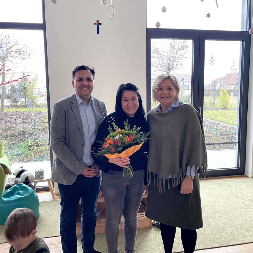 Drei Erwachsene und ein Kind posieren für ein Foto in einem Raum mit großen Fenstern und einem Baum draußen. Eine Frau hält einen Blumenstrauß.