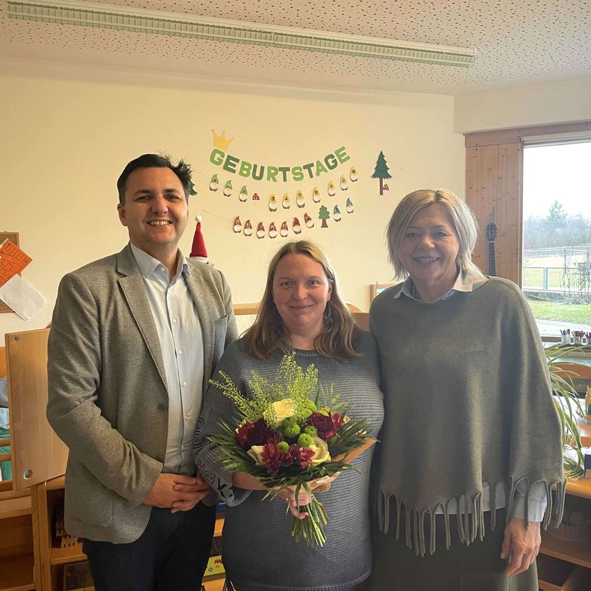 Drei Menschen stehen nebeneinander und lächeln in einem Raum mit einer weißen Wand und einem Banner, auf dem 'Geburtstag' steht. Die Frau in der Mitte hält einen Blumenstrauß. Hinter ihnen befindet sich ein Fenster mit Blick auf Bäume und eine Gitarre.