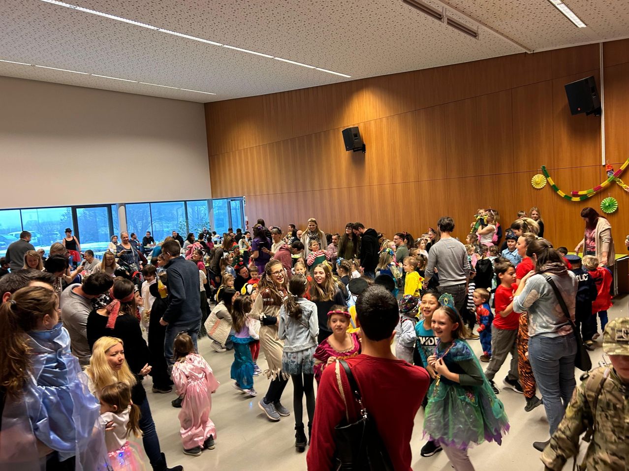 Eine große Gruppe von Kindern und Erwachsenen ist drinnen versammelt, wahrscheinlich für eine Kostümparty. Sie sind in verschiedenen Kostümen gekleidet und einige halten Taschen. Ein Mann in einem roten Shirt steht vor ihnen. Der Raum hat Holzwände und Glasfenster.